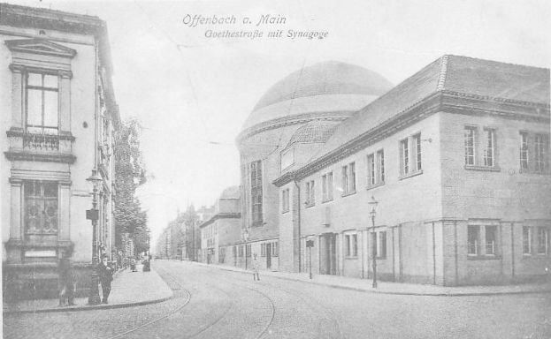 Ein Schwarz-Weiß-Foto einer Straße namens Goethestraße in Offenbach am Main, das Gebäude, Straßenlaternen, Bäume und Passanten zeigt, mit der Aufschrift "Offenbach a Main, Goethestraße mit Synagoge" oben.