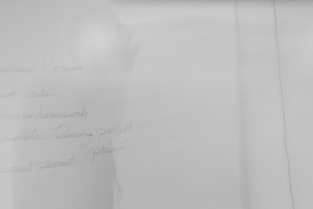 Ein Hund sitzt auf einem Bett neben einer Puppe und schaut aus einem Fenster mit einem Vorhang auf der linken Seite, während ein Auto auf der Straße draußen neben einem Baum geparkt ist, mit einer Sofa rechts, das mit Decken bedeckt ist.