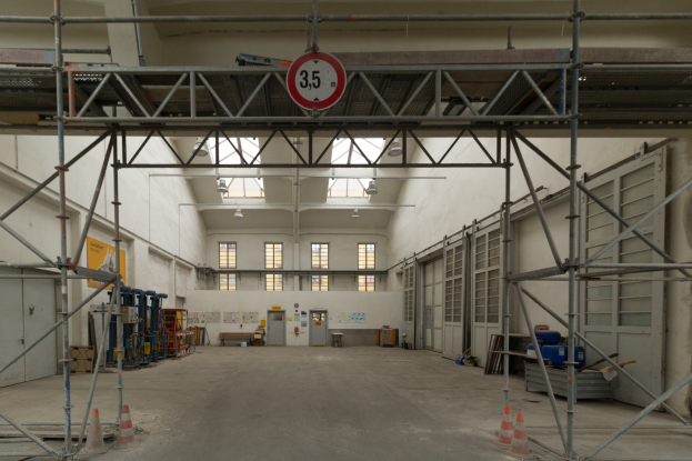 Ein großes Lager mit ausgedehnten Gerüsten, Verkehrskegeln auf dem Boden, Metallstangen, ein Schild, verschiedene Maschinen, eine Tür, Fenster, eine Wand und ein Dach mit Deckenbeleuchtung.