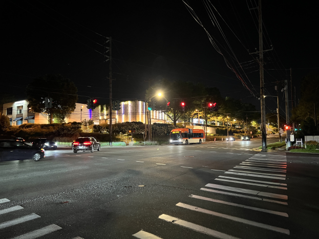 Eine nächtliche Stadtstraße mit fahrenden Autos und einem Bus, beleuchtet von Straßenlaternen und Ampeln, gesäumt von Gebäuden, Bäumen und Gras, unter einem dunklen Himmel.