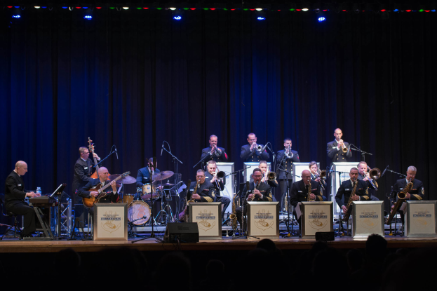 Eine Gruppe von Musikern auf der Bühne, die Instrumente spielen, mit Mikrofonen und Ständern, Schilder mit der Aufschrift "The New Orleans Jazz Band", ein Vorhang im Hintergrund und ein Publikum darunter unter Bühnenlichtern.