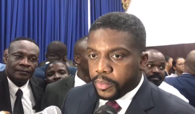 Ein Mann im Anzug und Krawatte spricht vor einer Gruppe von Menschen in ein Mikrofon, mit Vorhängen und einer Wand im Hintergrund, als ob er auf eine lebenslange Haftstrafe reagiert.