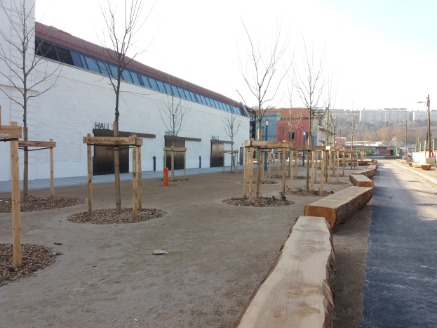 Ein Spielplatz mit Holzbänken und Bäumen vor einem Gebäude, umgeben von Straßenlaternen, Autos und Gebäuden unter einem klaren blauen Himmel.