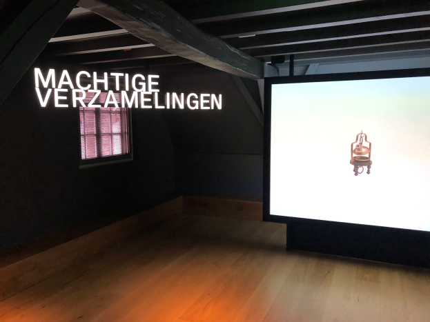 Installationsansicht der Ausstellung "Machtige Verzamelingen" im Museum of Modern Art in Berlin, mit einem großen Bildschirm mit Text an der Wand, einem Fenster links und einem Teil der Decke oben.