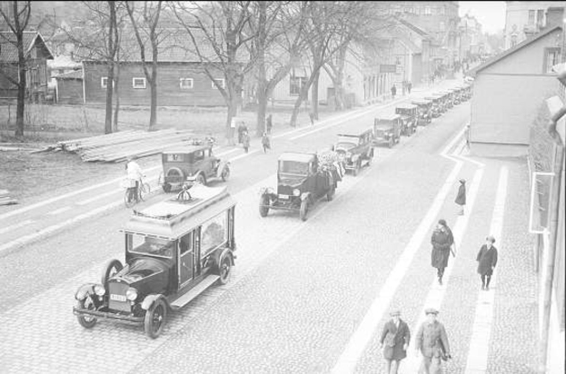 Schwarze und weiße Straßenszene in Berlin aus den 1930er-1940er Jahren, die vintage Autos, Fußgänger, Gebäude mit Fenstern, Bäume und einen klaren Himmel zeigt.