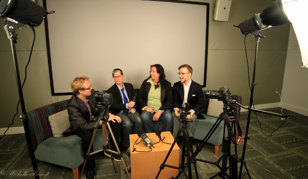 Eine Gruppe von drei Personen sitzt auf einer Couch und gibt ein Interview. Links sitzt eine Person in einem schwarzen Mantel und Hose, in der Mitte eine Person in einem Mantel und blauer Krawatte mit einem Mikrofon und rechts eine strahlende Person. Auf dem Tisch in der Mitte steht ein Mikrofonrekorder, Kameras sind auf der rechten und linken Seite und große Scheinwerfer sind vorhanden, sowie eine Projektionswand im Hintergrund.