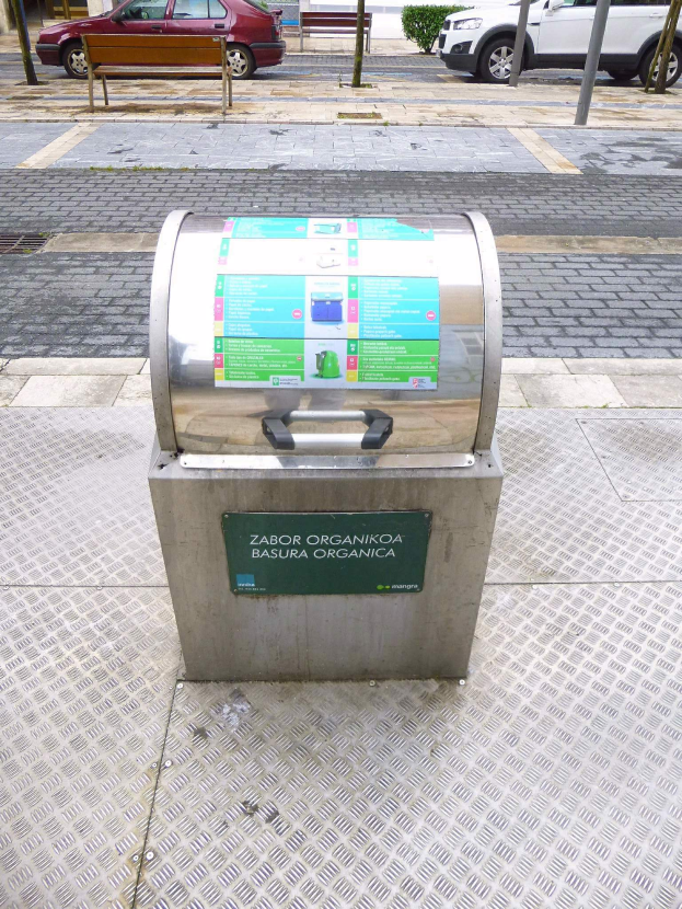 Ein Metallmüllbehälter am Straßenrand mit Fahrzeugen, einer Bank, Pfählen, Pflanzen, Bäumen und Gebäuden im Hintergrund.