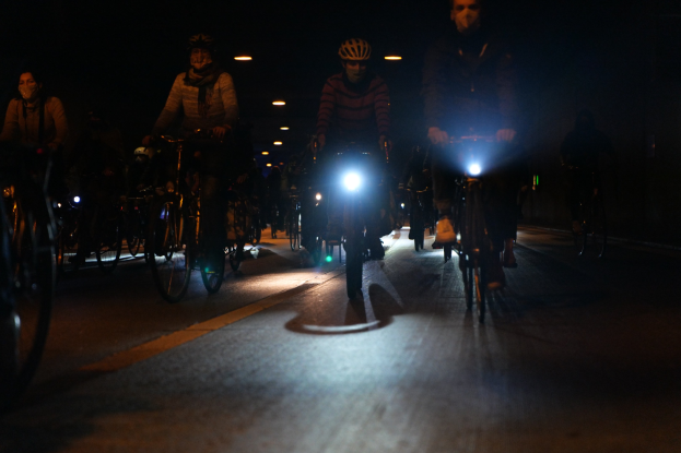 Eine Gruppe von Menschen, die nachts auf Fahrrädern durch eine Stadtstraße fahren, tragen Helme und werden von Stadtlichtern beleuchtet.