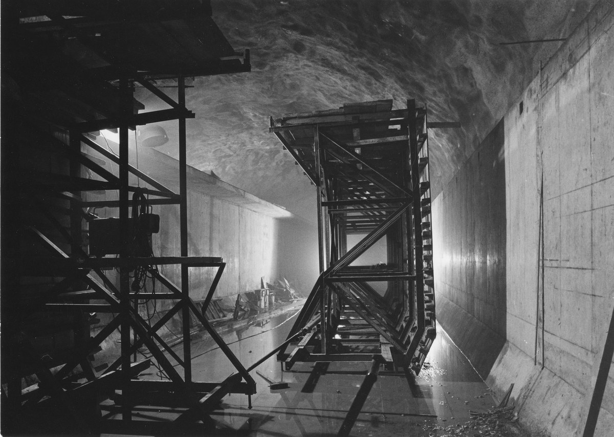 Schwarzes und weißes Foto eines Tunnels mit einer zentralen Metallstruktur, Metallstangen auf der linken Seite, einer Wand auf der rechten Seite und Text unten.