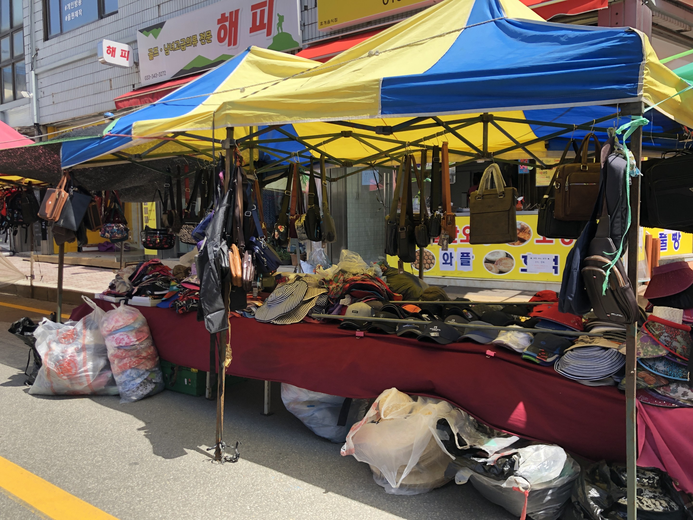 Ein Straßenmarkt mit Zelten, die Taschen, Hüte und andere Gegenstände ausstellen, vor Gebäuden mit Fenstern und Texttafeln.