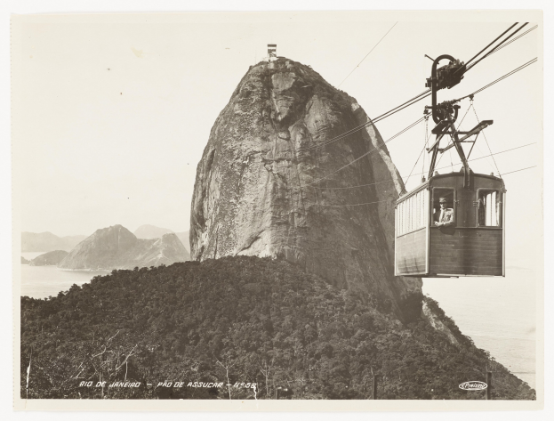 Schwarz-weißes Foto einer Seilbahn, die einen Berg hinauffährt, mit einer Person in der Gondel, umgeben von Bäumen, Hügeln und Wasser, mit Text am unteren Rand.