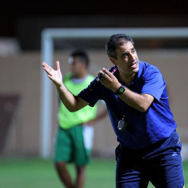 Ein Mann in einem blauen T-Shirt und blauen Hosen steht auf einem Fußballfeld und hält ein Mikrofon in der Hand, während er auf seine Spieler zeigt. Im Hintergrund ist eine Person in einem grünen Kleid, ein Tor und eine Wand zu sehen.