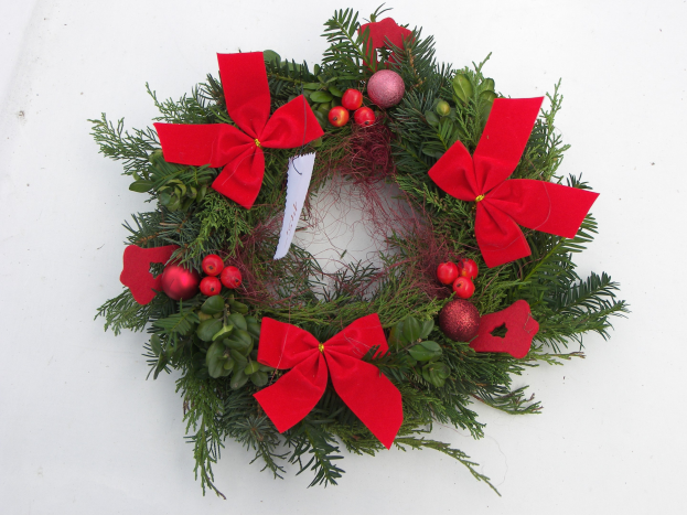 Ein festlicher Adventskranz mit roten Schleifen und Schmuck auf einer weißen Oberfläche, geschmückt mit üppigen grünen Blättern und leuchtend roten Bändern.