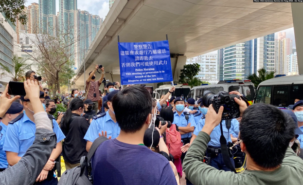 Eine Gruppe von Menschen steht vor einer Menge mit Kameras und Handys, mit einem Banner mit der Aufschrift "Hong Kong Police Wants to Stop the Violence Against Women", Fahrzeugen, Bäumen, Gebäuden und einem bewölkten Himmel im Hintergrund.