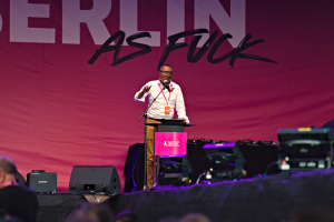 Ein Mann in einem weißen Hemd und braunen Hosen steht auf einer Bühne und hält ein Mikrofon in der Hand, mit einem pinken Banner im Hintergrund, das 'Berlin as Fuck' lautet und Lautsprecher und andere Gegenstände auf der Bühne.