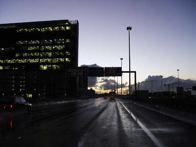 Ein durchsichtiges Gebäude, das mit Lichtern gefüllt ist, mit mehreren Strommasten und einer Straße mit ein paar Fahrzeugen auf der rechten Seite.