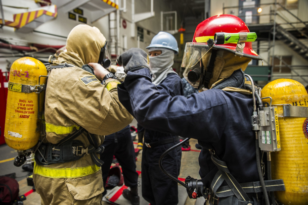 Feuerwehrleute in Schutzausrüstung mit Helmen, Handschuhen und Sauerstoffflaschen stehen zusammen bei Taschen, Treppen, Geländern und Lampen im Hintergrund.