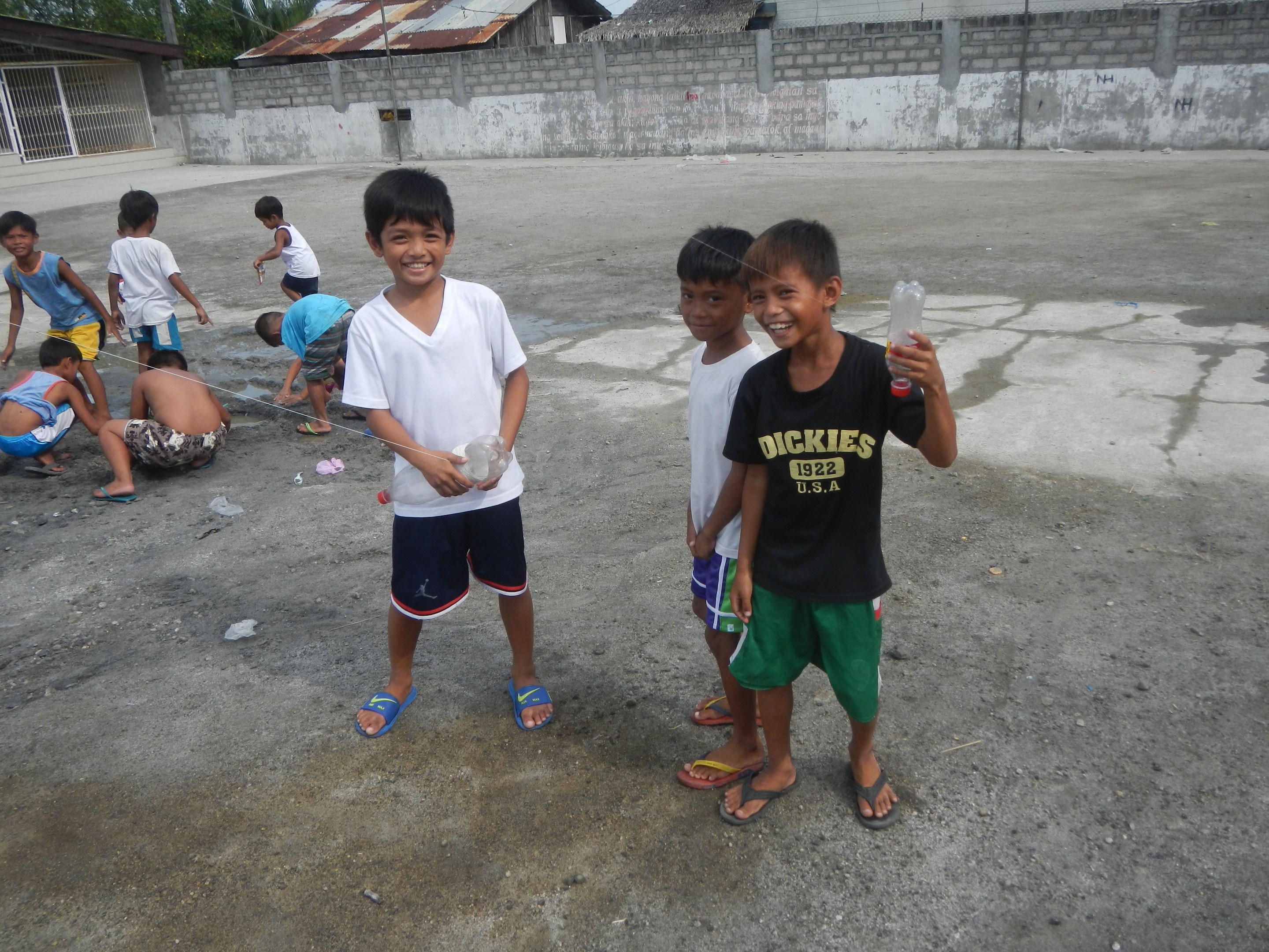 Gruppe junger Jungs, die in einer Parkbucht stehen und Flaschen halten, mit Häusern, Bäumen und Pfählen im Hintergrund und einem klaren blauen Himmel.