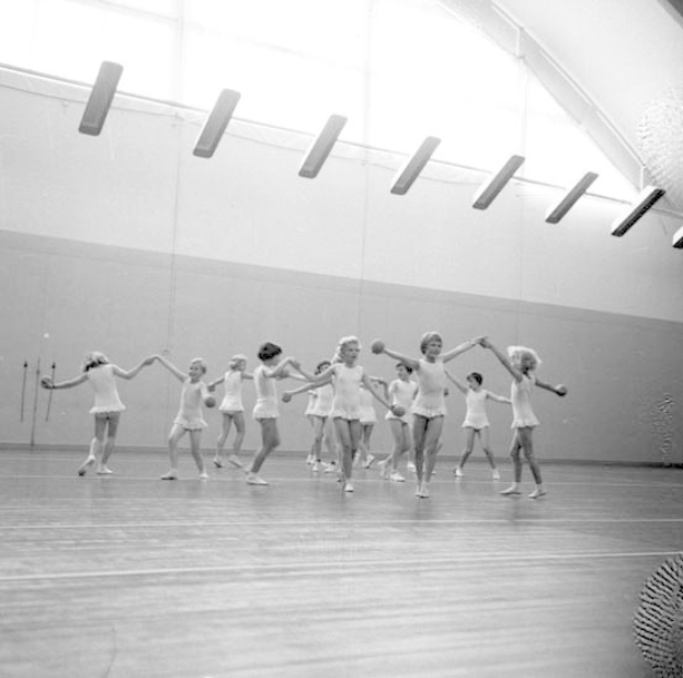 Schwarz-weiß-Bild einer Gruppe junger Mädchen, die in einem Studio tanzen und sich an den Händen halten, mit einem Ball auf der rechten Seite und Lampen an der Decke.