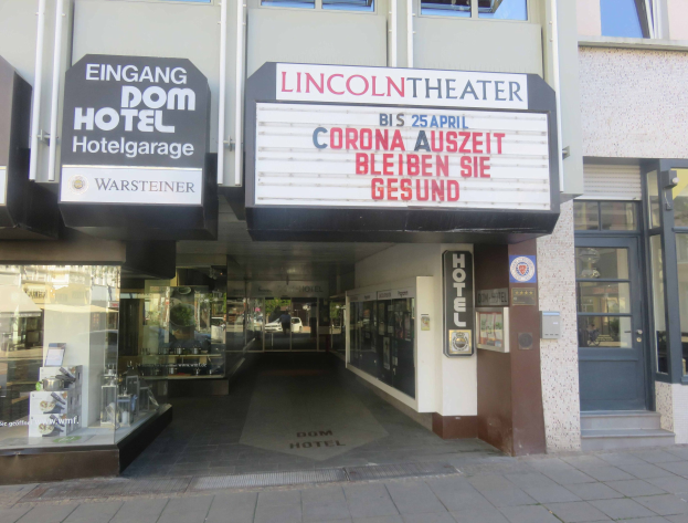 Das Lincoln Theater in Berlin, Deutschland, mit Glasfenstern und -türen und einer Tafel mit Text sowie einem Inneren, das eine belebte Stadtlandschaft suggeriert.