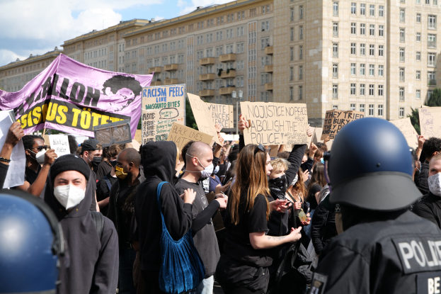 Eine Gruppe von maskierten Menschen mit Schildern während einer Demonstration, mit zwei Polizisten in Helmen auf der rechten Seite, vor einem Hintergrund aus Bäumen und einem bewölkten Himmel.