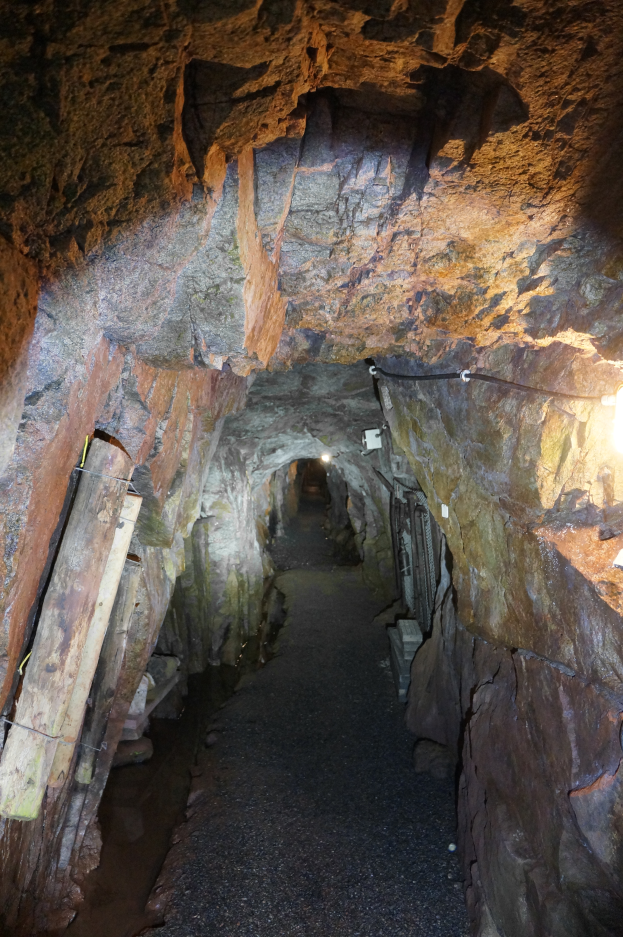 Ein langer, schwach beleuchteter Stollentunnel mit gewundenem Pfad, der zu einem Licht am Ende führt, umgeben von hölzernen Stützen.