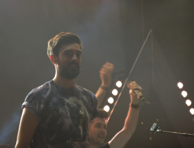 Ein Mann in einem T-Shirt steht und lacht, mit einem anderen lachenden Mann, Lampen, Rauch, einem Mikrofon und anderer Ausrüstung im Hintergrund.