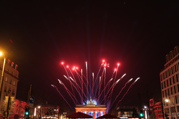 Eine Stadtstraße mit dem beleuchteten Brandenburger Tor in Berlin, Deutschland, nachts, umgeben von Gebäuden, Bäumen und Menschen, mit Feuerwerk am Himmel.