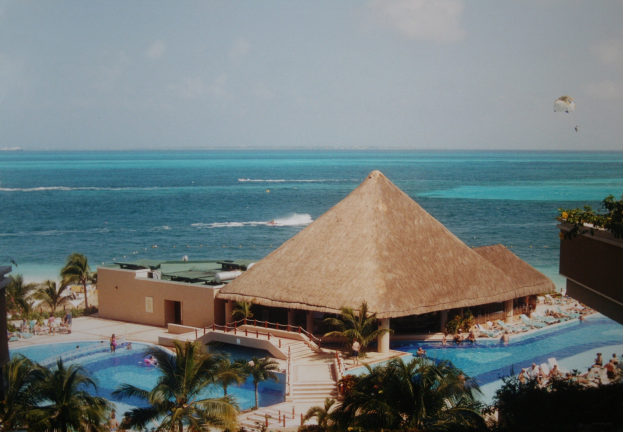 Resort mit einem gro├čen Schwimmbad umgeben von Palmen, Hütten und Menschen, mit einem Ozean im Hintergrund unter einem bew├Âlktem Himmel und einem Fallschirm in der Luft.