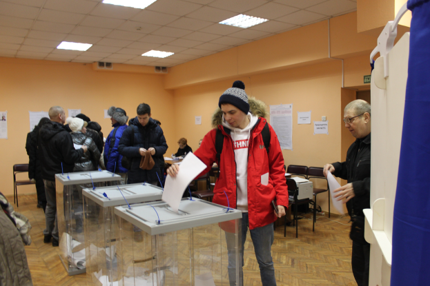 Eine Gruppe von Menschen steht um eine Wahlkabine in einer Wahllokal, einige halten Papiere, mit einem blauen Vorhang und einem weißen Objekt auf der rechten Seite, Stülhle, Tische, Papiere an der Wand und Deckenlampen im Hintergrund.