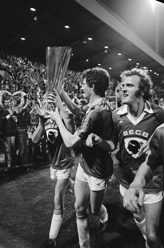 Eine Gruppe von Männern in Schwarz-Weiß-Kleidung hält einen Pokal auf einem Fußballfeld hoch und feiert ihren FA-Cup-Finalsieg, während eine Menge hinter einem Zaun und von Deckenlampen beleuchtet zusieht.