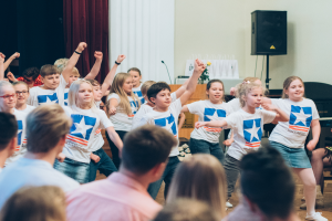 Eine Gruppe von Kindern in weißen Hemden und blauen Shorts, die vor einem sitzenden Publikum tanzen, mit einer Vorhangwand, Lautsprecherstativ und einem Tisch mit Mikrofon im Hintergrund.