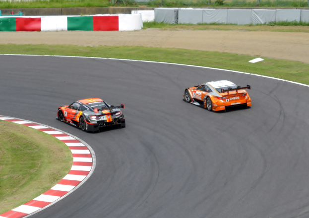 Zwei orangen Sportwagen auf einer Rennstrecke mit Gras auf beiden Seiten, einem Zaun, Pfosten, Bäumen und einem klaren blauen Himmel im Hintergrund.