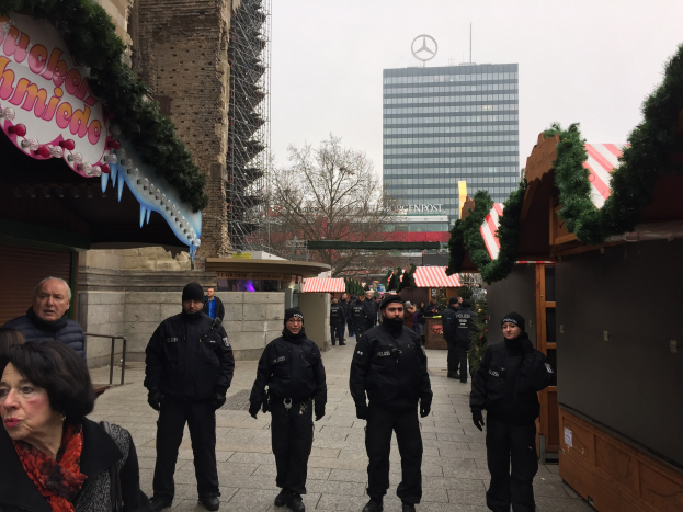 Polizeibeamte in schwarzen Uniformen stehen vor einem Berliner Weihnachtsmarkt mit festlicher Dekoration, Gebäuden, Bäumen und einem klaren blauen Himmel im Hintergrund; ein Schild mit Text ist links sichtbar.
