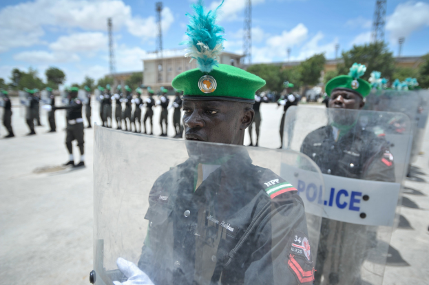 Eine Gruppe nigerianischer Polizeibeamter steht vor einer Reihe uniformierter Personen mit Schilden, mit Bäumen, Türmen, Gebäuden und einem bewölkten Himmel im Hintergrund.