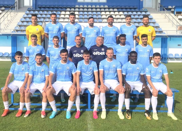 Gruppe von Männern auf einem Fußballfeld, einige sitzen auf Bänken und andere stehen auf dem Rasen, mit Stadion-Sitzplätzen und Gebäude-Strukturen im Hintergrund.