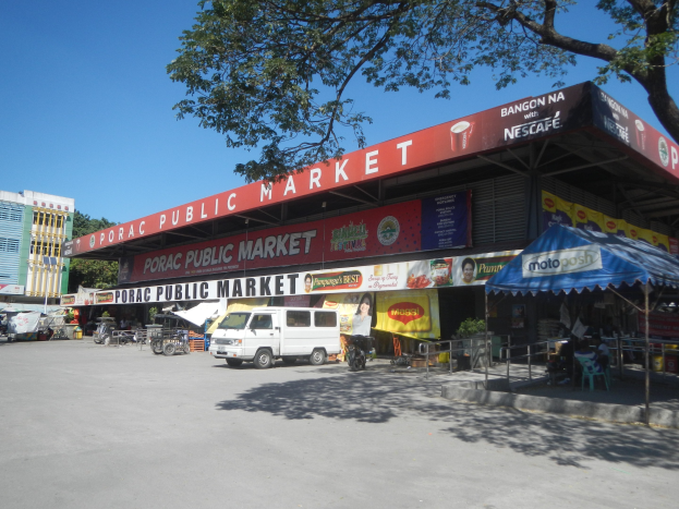 Ein öffentlicher Markt mit einem vor ihm geparkten Van, umgeben von Gebäuden, Geschäften, Schildern, Fahrzeugen auf der Straße, Menschen unter einem Zelt sitzend, Geländern, Bäumen und einem klaren blauen Himmel.