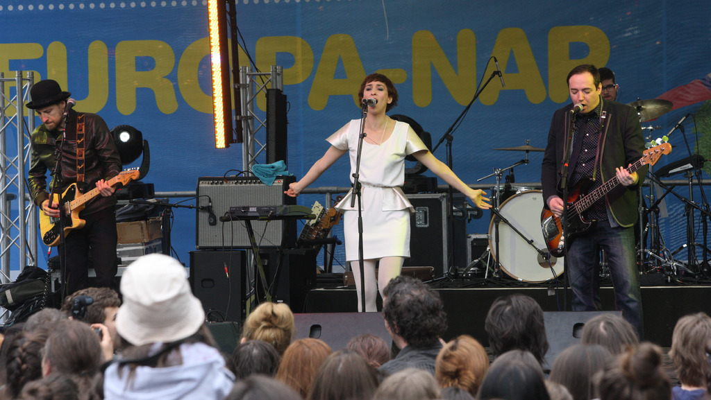 Eine Bühne mit vier Musikern: eine Frau in einem weißen Kleid singt in der Mitte, zwei Männer spielen Gitarren auf der linken und rechten Seite, eine Menge vorne und Lautsprecher an einer Wand im Hintergrund.