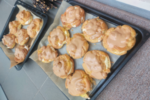 Ein Tablett mit verschiedenen Danish-Pasteten in hellgoldener Farbe und flaky Textur, einige mit Puderzucker bestäubt, liegt auf einer Theke neben einem offenen Fenster, gebadet im warmen Licht.