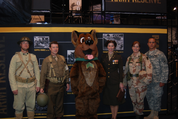 Personen stehen im Vordergrund, eine in einem Cartoon-Kostüm, mit Bannern und einem stangenähnlichen Objekt im Hintergrund.