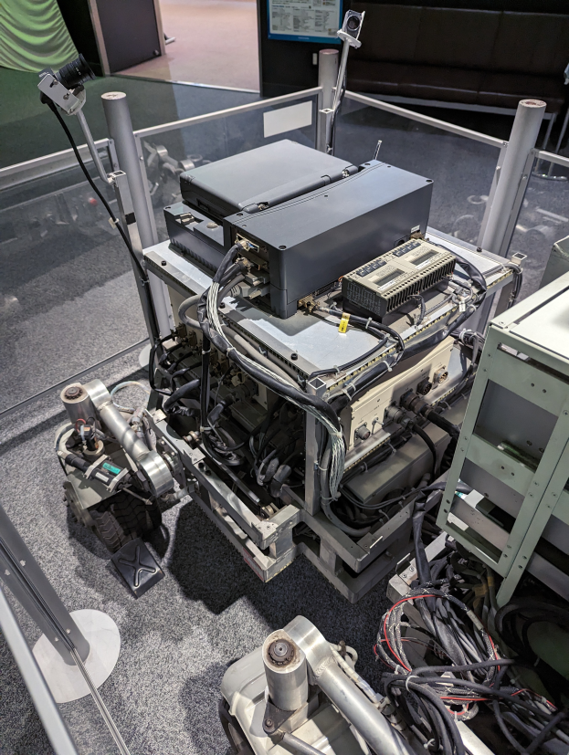 Ein automatisierter Apparat mit verschiedenen Komponenten und Kabeln steht auf einem Tisch neben einer Glaswand, mit einer Person rechts daneben; Sofas und ein Bildschirm sind im Hintergrund zu sehen.