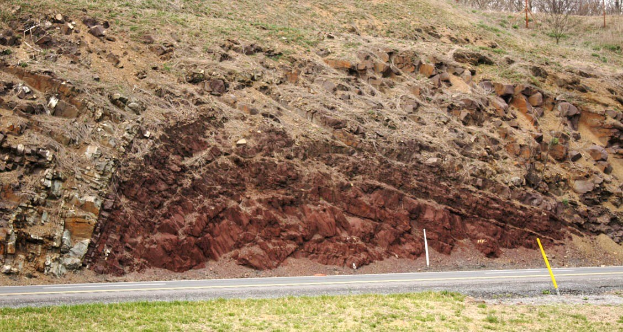 Großer Erdrutsch an der Seite einer Straße, mit Gras am Boden, einem Hügel in der Mitte, Bäumen und Himmel im Hintergrund.