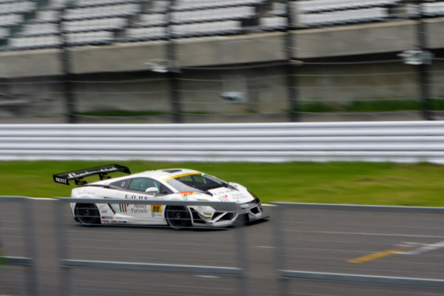 Weißer Sportwagen mit hoher Geschwindigkeit auf einer Rennstrecke, umgeben von einem Zaun und Gras, mit einem unscharfen Hintergrund.
