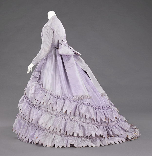 Eine Schaufensterpuppe in einem lila Ballkleid mit engem Oberteil, vollem Rock und Rüschen an den langen Ärmeln vor einem grauen Hintergrund.