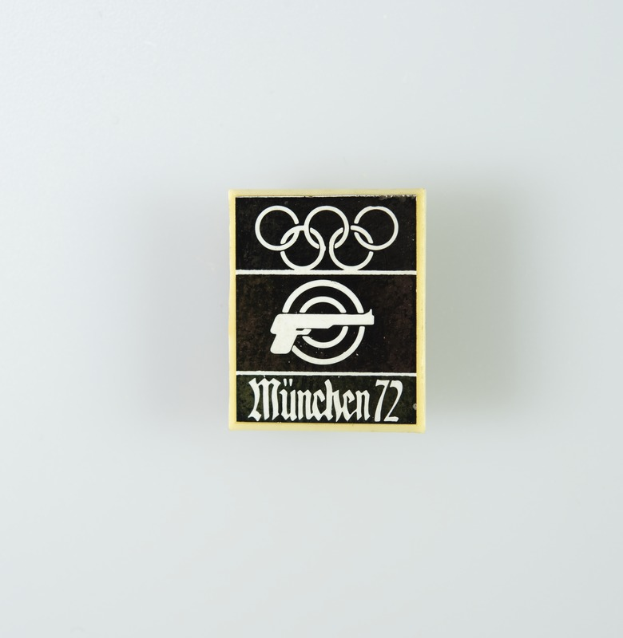 Ein schwarzes und weißes Logo mit einem Kreis und einer weißen Silhouette einer Person mit ausgestreckten Armen sowie der schwarzen Schrift 'München 1972'.