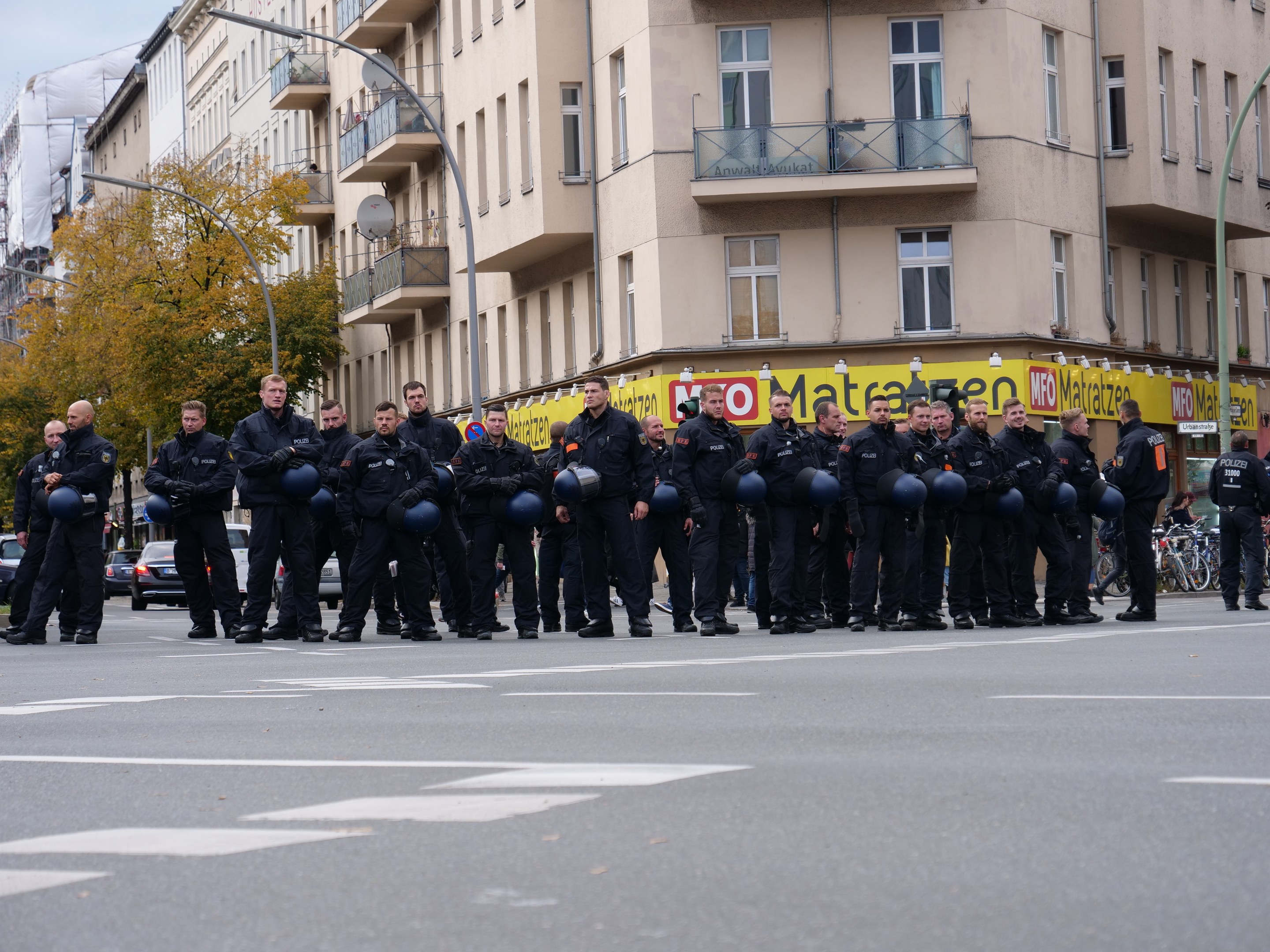 Eine Gruppe von Polizisten in schwarzen Uniformen mit blauen Helmen steht auf einer Straße, die von Laternenmasten, Bäumen und Glasfenstern gesäumt ist, unter einem klaren blauen Himmel.