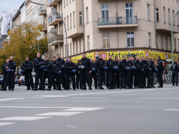 Eine Gruppe von Polizisten in schwarzen Uniformen mit blauen Helmen steht auf einer Straße, die von Laternenmasten, Bäumen und Glasfenstern gesäumt ist, unter einem klaren blauen Himmel.