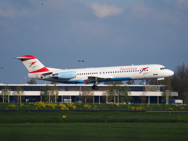 Ein Airbus A320-200 der Austrian Airlines beim Start am Frankfurter Flughafen, umgeben von grünem Gras, blühenden Pflanzen, Bäumen, Laternenmasten und einem Gebäude im Hintergrund unter einem bewölkten Himmel.