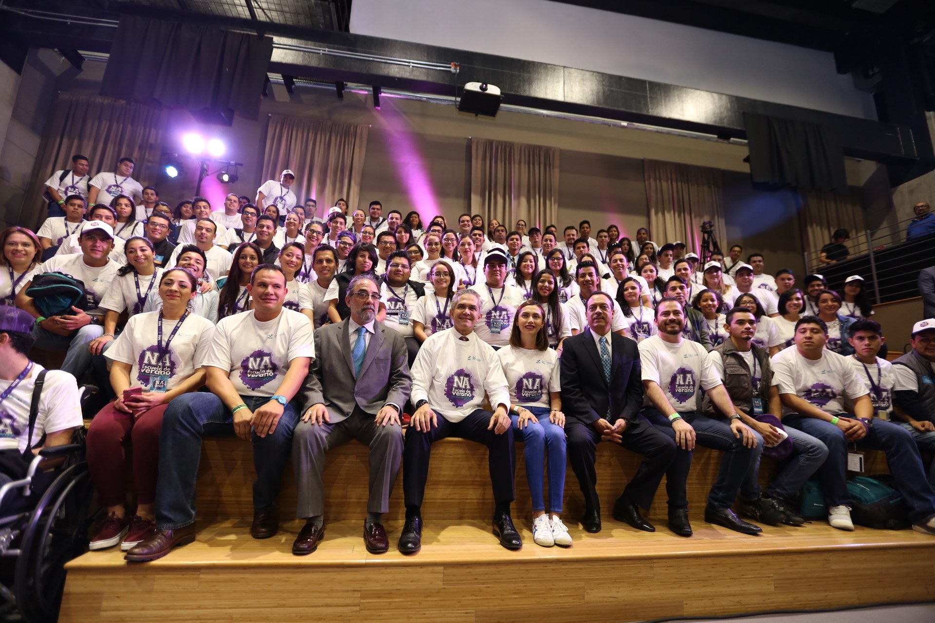 Eine Gruppe von Menschen sitzt auf einer Bühne mit einer Person im Rollstuhl links daneben, vor einem Vorhang, Scheinwerfern und einer Wand auf einer Veranstaltung, die wahrscheinlich mit dem Nationalen Frauenbasketballverband in Verbindung steht.