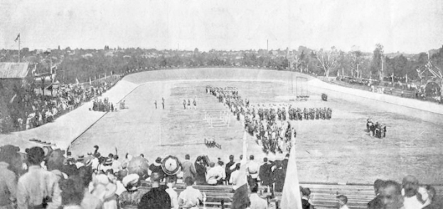 Schwarzes und weißes Foto einer Menge, die ein Pferderennen in einem Stadion beobachtet, mit Zuschauern auf Bänken und Rasen, einige halten Fahnen, und Reiter auf Pferden vor einem Hintergrund aus Bäumen und Himmel.